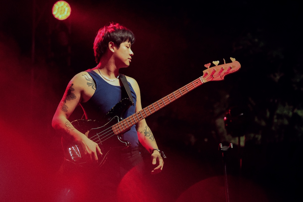 Live performance shot of musician Don Glori playing a bass guitar on stage under warm red concert lighting.